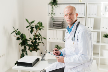 Aufkleber - Handsome young doctor in whitecoat sitting on desk while making medical notes