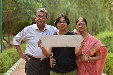 Wall Mural - Senior Bengali Indian couple with her young daughter holding a blank placard which can be used to write a message in a park in New Delhi, India