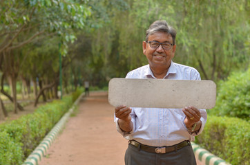 Senior Bengali Indian man holding a blank placard which can be used for quotes in a park in New Delhi, India. Concept motivational