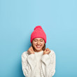© wayhome.studio  - Vertical image of joyful young girl feels overjoyed, raises clenched fists, being in good mood, wears white sweater and pink hat, dressed in warm clothes during cold autumn day, isolated on blue wall