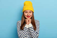 Image Of Tender Good Looking Dark Haired Woman Stands Against Blue Background In Yellow Hat And Striped Sweater, Wears Red Lipstick, Makeup, Looks Staightly At Camera. People, Beauty, Fashion