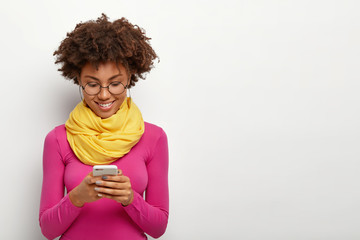 Horizontal shot of pleased dark skinned female holds modern mobile phone, focused into display, wears pink turtleneck, sends pictures online, browses internet page, stands against white wall