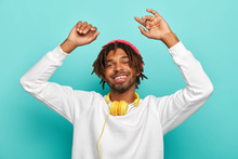 Horizontal View Of Optimistic Relaxed Afro American Man With Dreads, Enjoys Beats While Listning Music Or New Track Via Headphones, Keeps Arms Raised, Has Alluring Smile, Dressed In White Sweater.