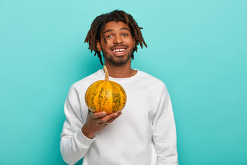 Wall Mural - Happy handsome young mixed race guy with dreads, holds little ripe yellow pumpkin, going to make diet salad, dressed in casual wear, carves vegetable for Halloween, isolated over blue background.