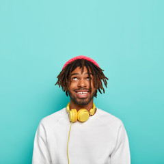 Wall Mural - Lively modern hipster with dreads, smiles happily, looks above, wears pink hat and white jumper, being amused by listening music via headphones, has happy mood, isolated over blue studio wall