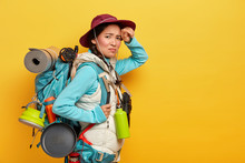 Tired Exhausted Female Tourist Walks Long Distance On Foot, Looks With Dissatisfaction At Camera, Stands Sideways Against Yellow Background, Carries Backpack With Personal Stuff. Traveling And Tourism
