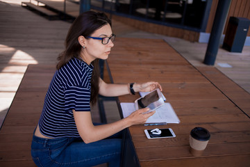 A serious brunette girl, freelancer, with large wrist watch, works at a wooden table on tablet, with gadgets and papers.