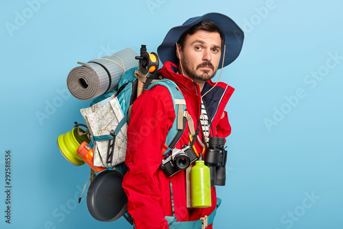 Fotomural Photo of active handsome man with mustache and bristle, carries tourist rucksack on back, walks in forest, has hiking trip, wears red jacket and hat, stands against blue background