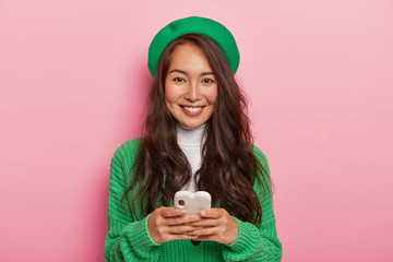 Wall Mural - Photo of brunette Korean female holds modern cellular, sends text messages, surfes internet, wears green beret and casual jumper, poses against pink background. Mixed race teenager with mobile phone