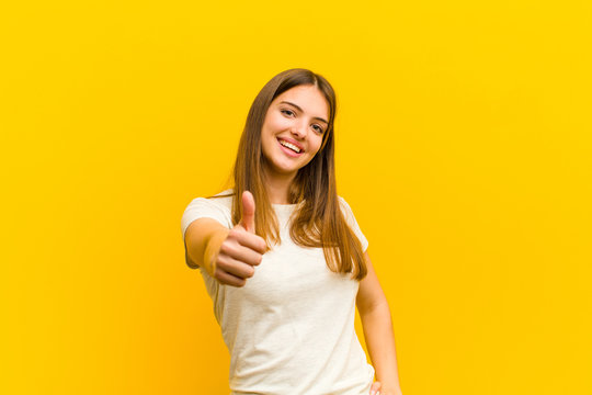 young pretty woman feeling proud, carefree, confident and happy, smiling positively with thumbs up against orange background