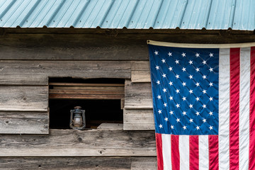 Rustic outside wall of wood building with green metal roof, open window with dusty lantern on windowsill, American flag hanging on wall