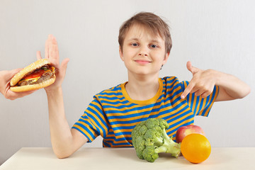 Little funny boy in a striped shirt at the table refuses hamburger in favor of fruit and vegetables on a white background