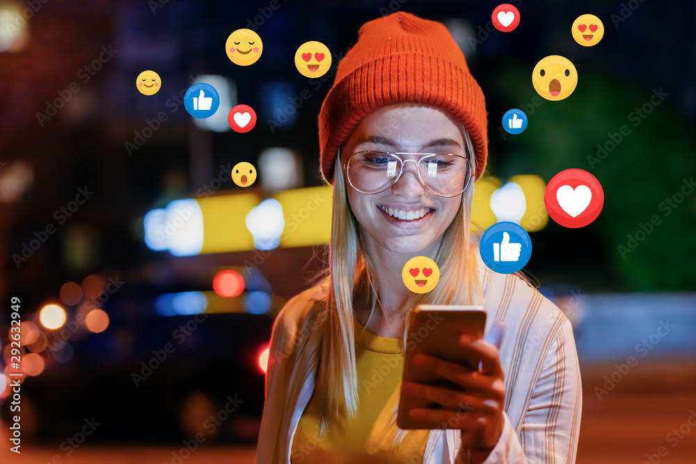 A beautiful young woman blogger, vlogger or influencer is receiving emoji and emoticon reactions in her mobile smart phone device while making a post, sharing or video logging on social media - obrazy, fototapety, plakaty 