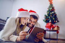Cute Caucasian Couple Lying On Stomach On The Floor With Santa Hats On Heads And Using Tablet For Online Shopping. Woman Holding Credit Card. Living Room Interior. In Background Is Christmas Tree.