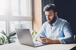 © foxyburrow - Young man sitting at window and working on laptop. Bearded man sitting at desktop with smartphone in his hands. Online education.