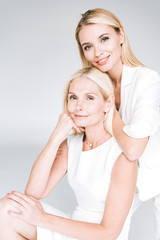 smiling blonde young daughter embracing mature mother in total white clothes isolated on grey