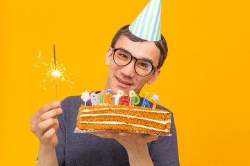 Wall Mural - Funny positive guy in glasses holds in his hands a homemade cake with the inscription happy birthday posing on a yellow background. Concept of holidays and anniversaries.