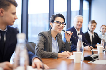 Wall Mural - Portrait of young businesswoman speaking to microphone during conference and smiling cheerfully while sitting in row with colleagues, copy space