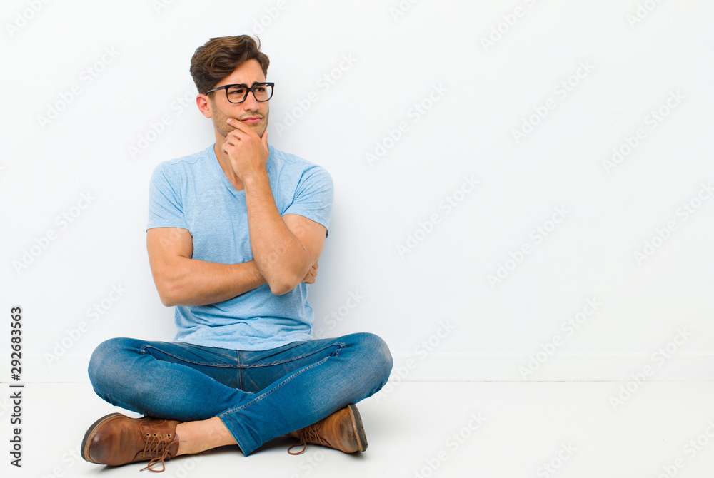 young handsome man thinking, feeling doubtful and confused, with different options, wondering which decision to make sitting on the floor - obrazy, fototapety, plakaty 