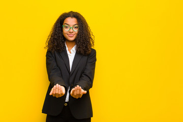 black business woman smiling happily with friendly, confident, positive look, offering and showing an object or concept against orange wall