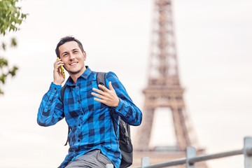 A smiling young man is chatting on the phone and gesticulating animatedly against the backdrop of the Eiffel tower in Paris.