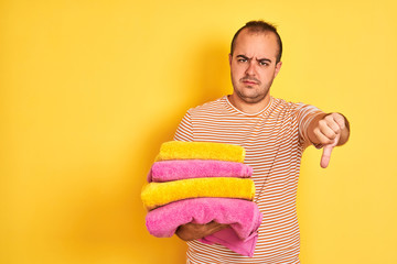 Canvas Print - Young shopkeeper man holding folded towels standing over isolated yellow background with angry face, negative sign showing dislike with thumbs down, rejection concept