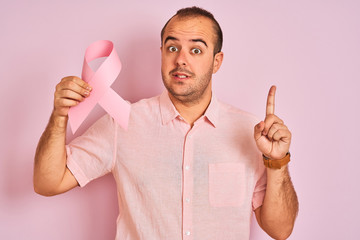 Young man holding cancer ribbon symbol standing over isolated pink background surprised with an idea or question pointing finger with happy face, number one