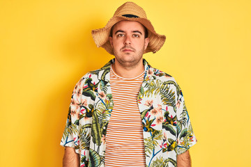 Sticker - Young man on vacation wearing summer shirt and hat over isolated yellow background with serious expression on face. Simple and natural looking at the camera.