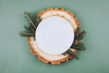 Festive Christmas natural style table setting with white plate on wood cut platters and fir tree branches on green blue background. Flat lay, top view, copy space