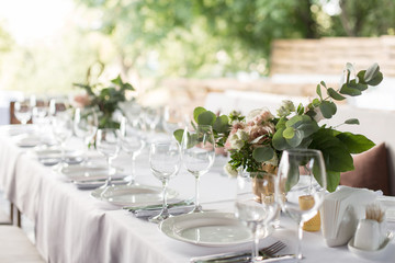 Wedding table setting decorated with fresh flowers in a brass vase. Wedding floristry. Banquet table for guests outdoors with a view of green nature. Bouquet with roses, eustoma and eucalyptus leaves