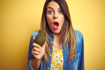 Poster - Young beautiful woman eating healthy avocado over yellow background scared in shock with a surprise face, afraid and excited with fear expression