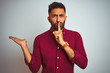 Young indian man wearing red elegant shirt standing over isolated grey background asking to be quiet with finger on lips pointing with hand to the side. Silence and secret concept.