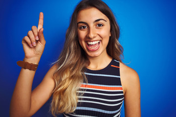 Wall Mural - Young beautiful woman wearing stripes t-shirt over blue isolated background surprised with an idea or question pointing finger with happy face, number one