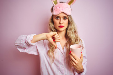 Wall Mural - Young woman wearing pajama and mask drinking a cup of coffee over pink isolated background with angry face, negative sign showing dislike with thumbs down, rejection concept