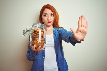 Sticker - Young beautiful redhead woman holding jar of cookies over white isolated background with open hand doing stop sign with serious and confident expression, defense gesture