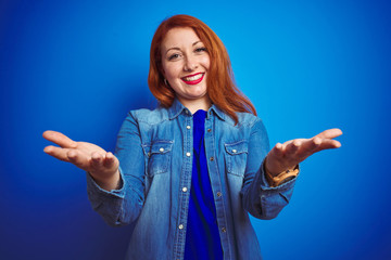 Sticker - Young beautiful redhead woman wearing denim shirt standing over blue isolated background smiling cheerful offering hands giving assistance and acceptance.