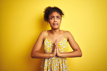 Sticker - African american woman wearing casual floral dress standing over isolated yellow background begging and praying with hands together with hope expression on face very emotional and worried