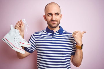 Sticker - Young man holding casual sneakers shoes over pink isolated background pointing and showing with thumb up to the side with happy face smiling