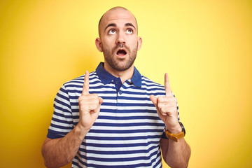 Sticker - Young bald man with beard wearing casual striped blue t-shirt over yellow isolated background amazed and surprised looking up and pointing with fingers and raised arms.