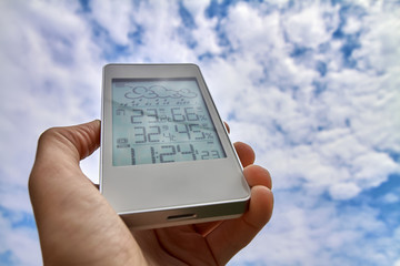 Wall Mural - Best personal weather station device with weather conditions inside and outside. A man holds a gadget in his hand on a background of sky and clouds