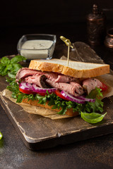 Wall Mural - Roast beef sandwich with tomato, onion, lettuce and mustard sauce on dark background. Delicious healthy lunch with meat, hearty food