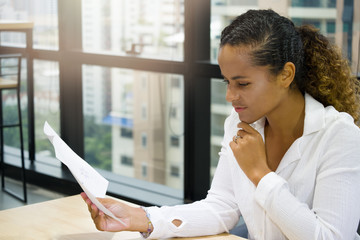 Adult American black woman sat at a desk full of documents and touching her chin with a finger while looking at a sheet of paper to check the accuracy before proposing to the client.