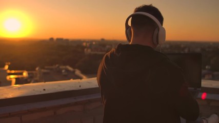 A man in headphones on the roof relaxes working remotely enjoying life despite a handsome kind of sipping beer and types on the keyboard.