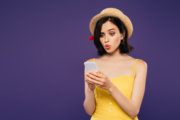 surprised pretty girl in straw hat using smartphone isolated on purple