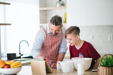 Wall Mural - Mature father and small son with tablet indoors in kitchen, making pancakes.