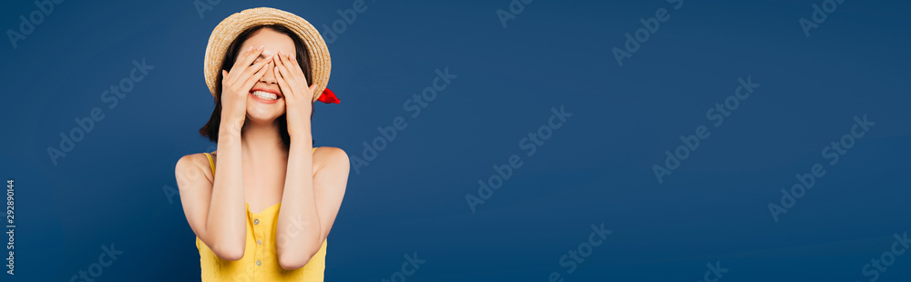 smiling girl in straw hat with hands on eyes isolated on blue - obrazy, fototapety, plakaty 