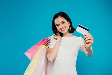 Wall Mural - smiling elegant woman in dress with shopping bags showing credit card isolated on blue