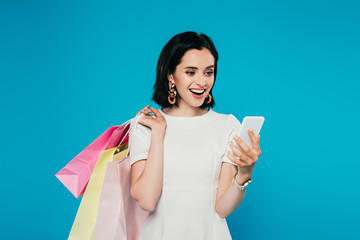 Wall Mural - happy elegant woman in dress with shopping bags holding smartphone isolated on blue