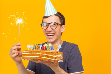 Wall Mural - Positive funny young asian guy with a cap and a burning candle and a homemade cake in his hands posing on a yellow background. Anniversary and birthday concept.