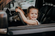 A little girl with long hair is sitting at the wheel of a car. Happy childhood, young driver, travel.